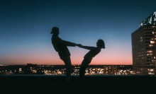 Two silhouetted people hold hands against a backdrop of twilight and city lights.