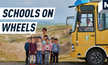 A group of Uzbek children and their teacher are posing for a picture. On the right, a yellow bus with solar panels on top is waiting for them. The driver inside looks at the camera, smiling. Behind them is a vast mountainous landscape. 