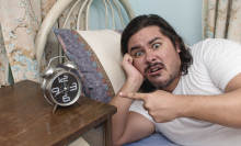 a man in bed looks irritated and confused about what his clock is telling him