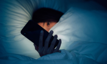 A young woman lies in bed, peeking at her phone from under the covers. 