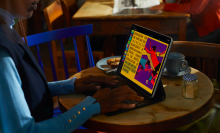 a woman in a blue vest and shirt types on an ipad pro's keyboard cover while sitting at a wooden table at a cafe