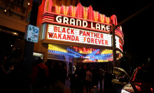 A glowing theater marquee sign reads, "Black Panther: Wakanda Forever". A crowd walks by under the sign. 