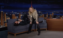 A blonde woman laughs while standing next to a man in a suit lying on his back on a talk show couch.