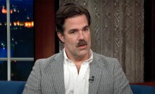 A man in a shirt and blazer sits in a talk show seat.