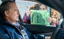 A man sits in his car with a woman showing him her child's drawing through the window. 