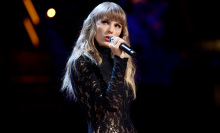 Taylor Swift performs onstage in a black sequin catsuit. Her hair is blonde and straight, hitting mid chest, and she has bangs. She is holding a microphone.