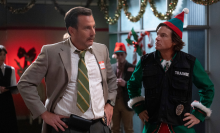 A man in a suit and a man in a Christmas elf costume talk. 