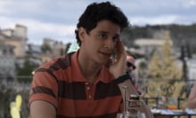 Albie (Adam DiMarco) sits at a table with one hand on his face and a faint smile on his mouth