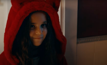 A young girl in a fluffy red coat smiles menacingly. 
