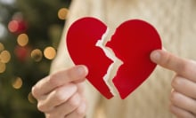 A person holds a paper heart with a rip down the middle, with a Christmas tree in the background. 