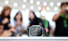 An apple watch rests on a white table in the middle of an Apple store. A group of customers are blurred out in the background.