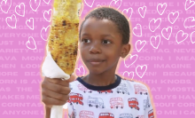 A photo of Tariq smirking at staring at an enormous ear of corn, which he clutches in his hand in the foreground.