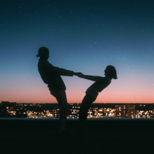 Two silhouetted people hold hands against a backdrop of twilight and city lights.