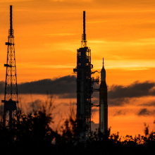 NASA's Space Launch System (SLS) rocket with the Orion spacecraft aboard — also known as Artemis I —sits on the launch pad on Aug. 31 ahead of its planned (and later cancelled) Sep. 3 launch. The sunset behind it is made of deep and bright oranges, and you can really only see the black outline of the rocket and system against it.