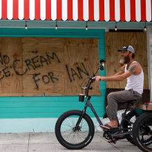 a man rides a bicycle in front of a boarded-up storefront upon which a shopkeeper has spray-painted "No ice cream for Ian."