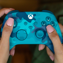 a close-up of a woman with light pink nail polish holding the new Xbox Wireless Controller – Mineral Camo Special Edition