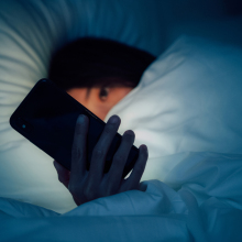 A young woman lies in bed, peeking at her phone from under the covers. 
