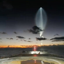 A plume of light curves around a distant rocket in the evening sky, seen from a droneship miles out at sea.