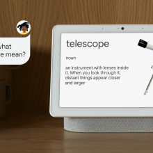 A Google smart screen device sits on a wooden table. On the screen is a kid-friendly definition of a telescope, which reads, "An instrument with lenses inside it. When you look through it, distant things appear closer and larger."
