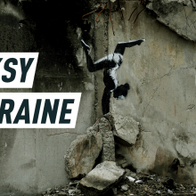 A mural painting depicts a female gymnast balancing a headstand on top of buildings ruins. Caption reads: "Banksy in Ukraine"