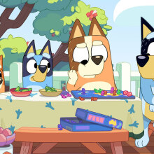 A family of cartoon dogs sit around a table outside.