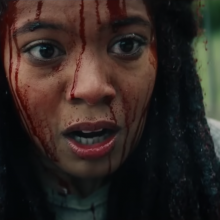 A close up of a Black woman in long locs. She looks surprised and her face dripping in blood.