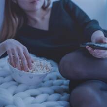 A woman reclines with her right hand in a bowl of popcorn and her left hand holding a remote control.
