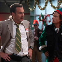 A man in a suit and a man in a Christmas elf costume talk. 