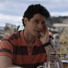 Albie (Adam DiMarco) sits at a table with one hand on his face and a faint smile on his mouth