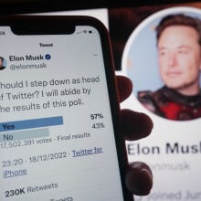 Elon Musk's Twitter poll asking if he should resign on a smartphone, in front of Musk's Twitter profile on a large screen.