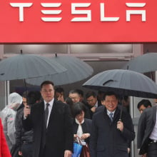 Elon Musk in the rain amid a crowd of people in business attire in front of a Tesla logo on a building
