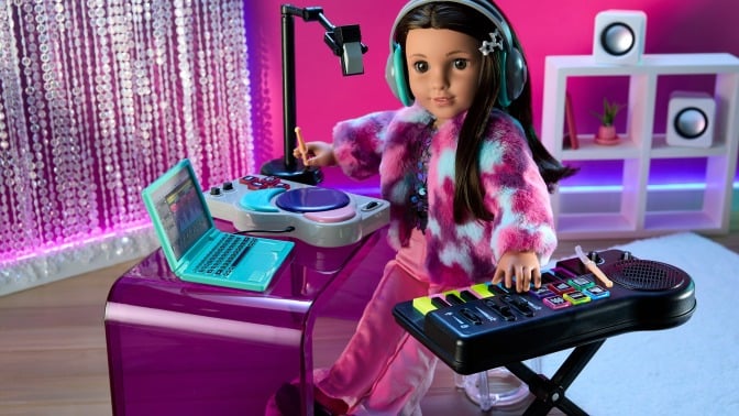 A doll is posed in a pink bedroom set. She is wearing headphones and sitting at a desk while playing an electric keyboard and looking at a laptop. 