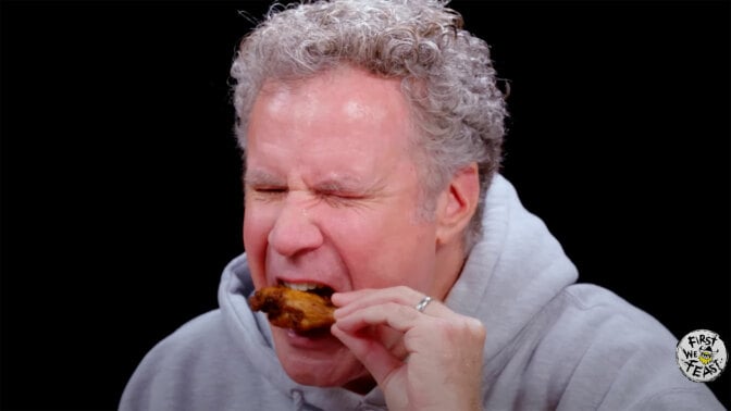 A man eats a chicken wing while closing his eyes in apparent discomfort.
