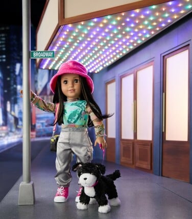 The Kavi doll is posed outside a miniature theater with a brightly lit marquee. She is holding the leash of a black and white dog and leaning on a lightpost. 