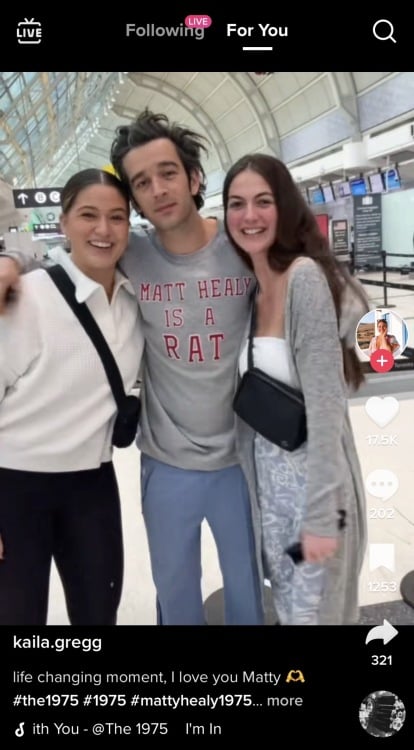 Three people: two young white women and Matty Healy in the middle with his arms around them, posing for a photo. He is wearing a grey sweatshirt that reads "Matt Healy is a rat."