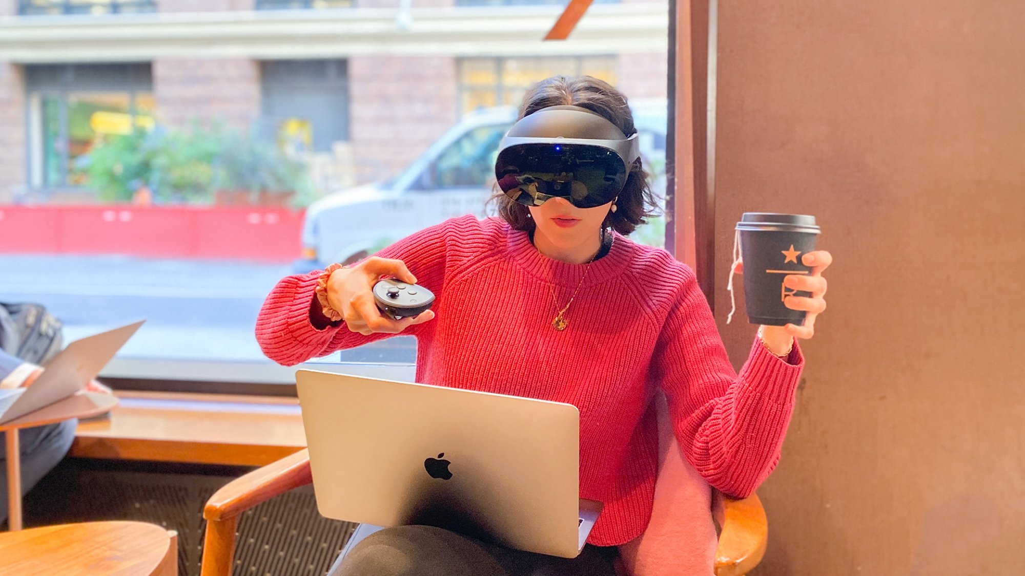 A woman sitting in a cafe with a laptop on her lap. She is wearing the Quest Pro and holds a to-go coffee cup in one hand and a controller in the other. 