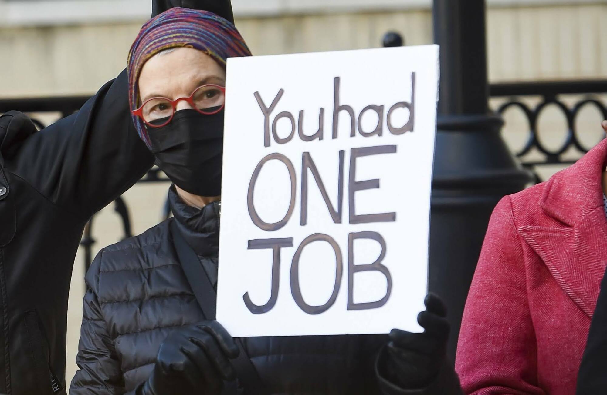 A woman bundled in a mask and padded jacket holds a white cardboard sign that reads, in black, "You had one job"
