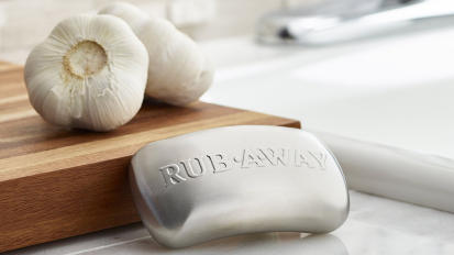 a rub away bar resting against a wood cutting board with two bulbs of garlic on it next to a kitchen sink