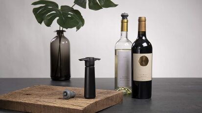 a vacu vin wine saver on a wooden board in front of two bottles of wine and a vase with two monstera leaves