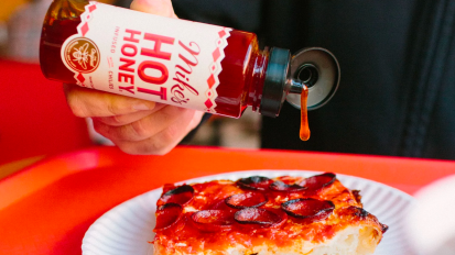 a close-up of a person pouring mike's hot honey on a slice of pepperoni pizza